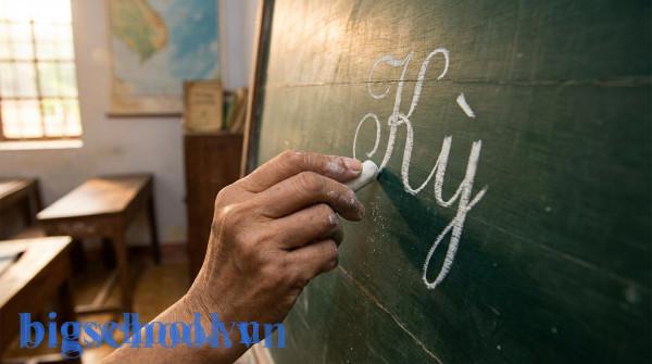 Cận cảnh bàn tay giáo viên Việt Nam đang viết chữ kỳ lên bảng xanh bằng phấn trắng.